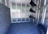 Interior of a metal trailer with silver walls and a blue speckled carpeted floor; wall-mounted black brackets and a small black storage bin in a corner, with CarolinaTrailerSales.com text visible along the bottom edge.