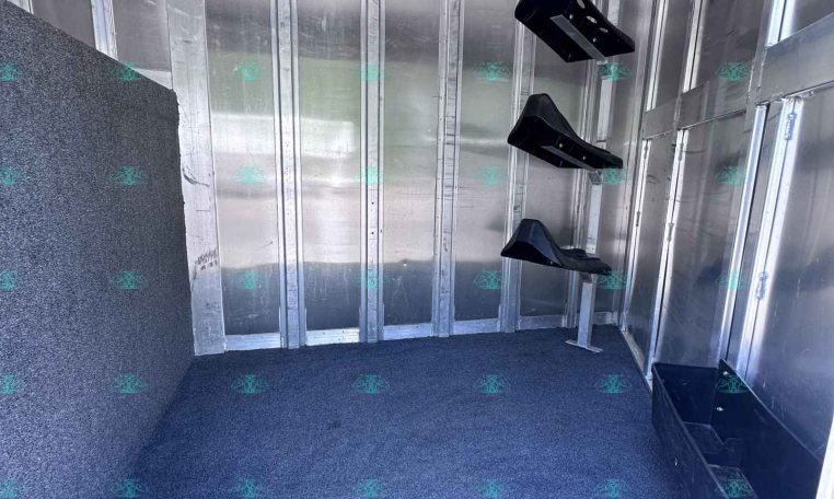 Interior of a metal trailer with silver walls and a blue speckled carpeted floor; wall-mounted black brackets and a small black storage bin in a corner, with CarolinaTrailerSales.com text visible along the bottom edge.
