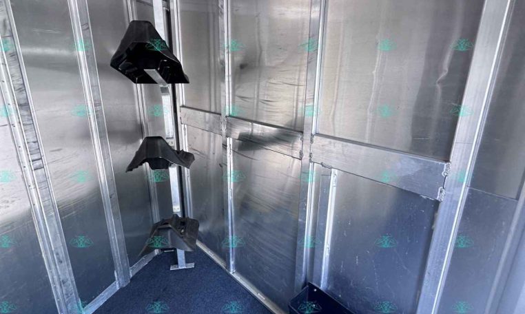 Interior of a silver cargo trailer with aluminum walls, vertical supports, and black seat frames mounted along the wall with a blue carpeted floor.