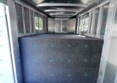 Empty aluminum trailer interior with a blue carpeted floor and side windows, viewed from the doorway at the rear area of the trailer.