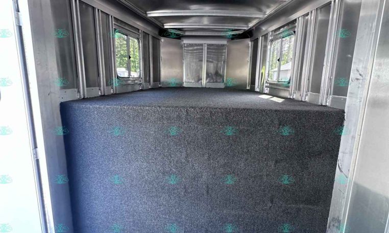Empty aluminum trailer interior with a blue carpeted floor and side windows, viewed from the doorway at the rear area of the trailer.
