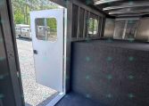 Interior view of a metallic trailer with a dark carpet, open door to outside, and windows along the side showing another trailer outside