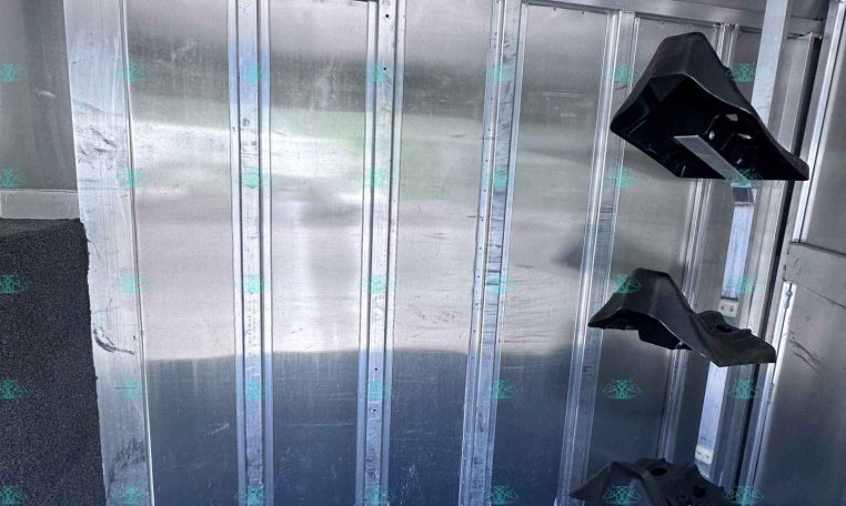 Interior of an aluminum trailer showing metal wall studs, a horizontal black plastic bracket, and a dark carpeted floor.