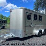 Front-right view of a large silver enclosed trailer with two side windows and tandem wheels, parked on gravel with trees and blue sky behind it.