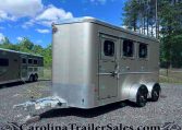 Front-right view of a large silver enclosed trailer with two side windows and tandem wheels, parked on gravel with trees and blue sky behind it.