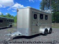 Front-right view of a large silver enclosed trailer with two side windows and tandem wheels, parked on gravel with trees and blue sky behind it.