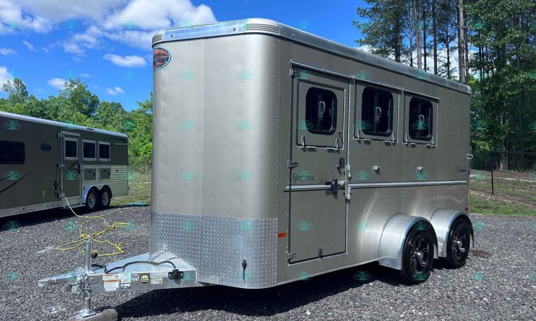 Front-right view of a large silver enclosed trailer with two side windows and tandem wheels, parked on gravel with trees and blue sky behind it.