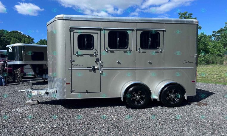 Silver aluminum horse trailer with three side windows parked on a gravel lot under a blue sky; branding reads 'Sportman' on the door side of the trailer.
