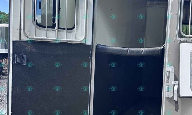 Open horse trailer door revealing interior with padded wall and metal framing on a gravel lot