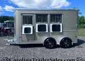 Silver horse trailer with three barred windows for horse stalls, parked on a gravel lot under a bright blue sky.