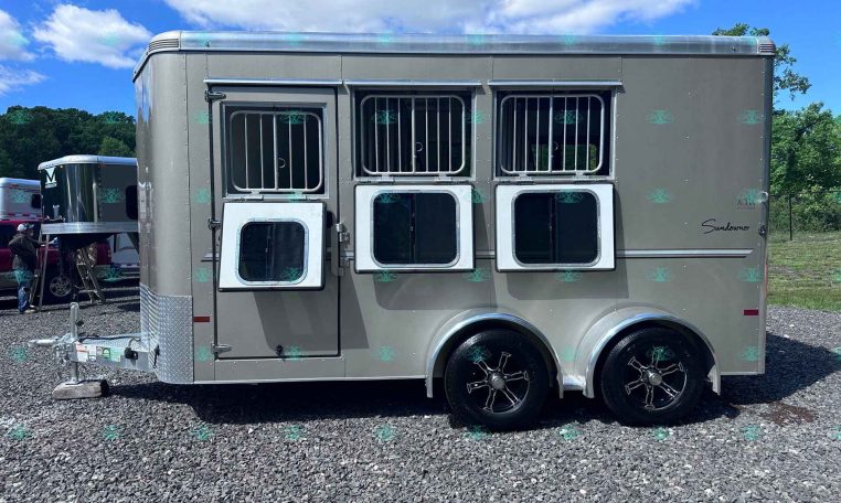 Silver horse trailer with three barred windows for horse stalls, parked on a gravel lot under a bright blue sky.