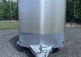 Front view of a silver enclosed cargo trailer with diamond-plate front, hitch attached, parked on a gravel lot with trees in the background.