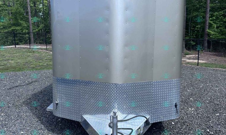 Front view of a silver enclosed cargo trailer with diamond-plate front, hitch attached, parked on a gravel lot with trees in the background.