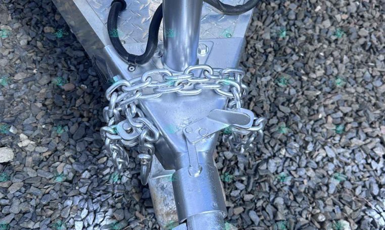 Close-up of a trailer hitch showing a silver diamond-plate trailer, safety chains, and a coupler at the tongue resting on gravel.