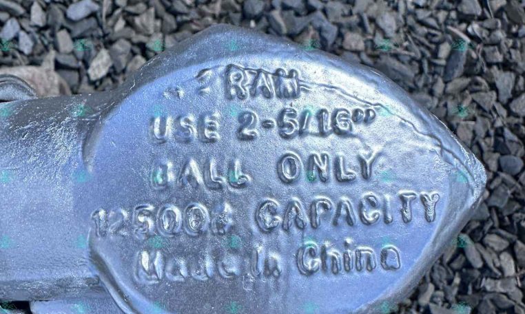 Close-up of a metallic trailer lock resting on gravel, showing stamped specifications like 2-5/16 inches and 1500 lb capacity on the body.