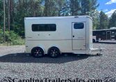 White horse trailer with two axles and large windows parked on a gravel lot, with a forest backdrop and another trailer in the background.
