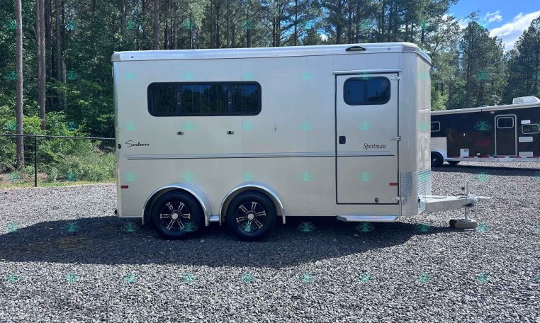 White horse trailer with two axles and large windows parked on a gravel lot, with a forest backdrop and another trailer in the background.