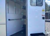 Open white horse trailer showing empty stalls, hanging hooks, and saddle racks inside with the door ajar on a sunny day.