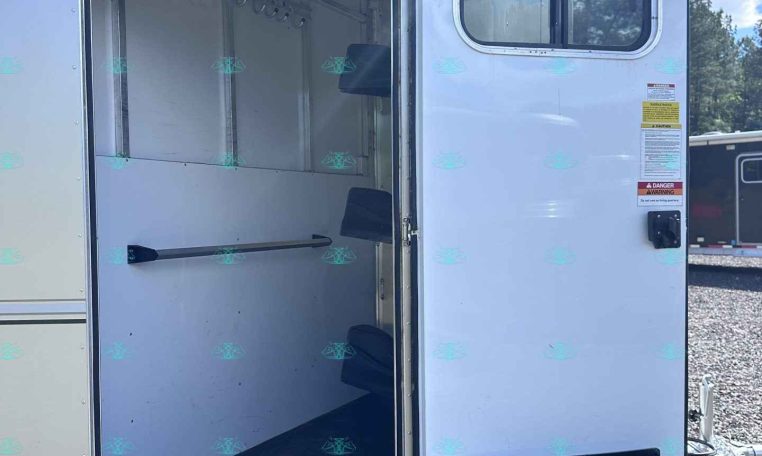 Open white horse trailer showing empty stalls, hanging hooks, and saddle racks inside with the door ajar on a sunny day.