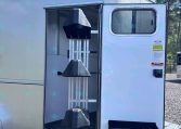 Open silver horse trailer with rear door and ramp down, showing interior stalls on a gravel lot with trees in the background