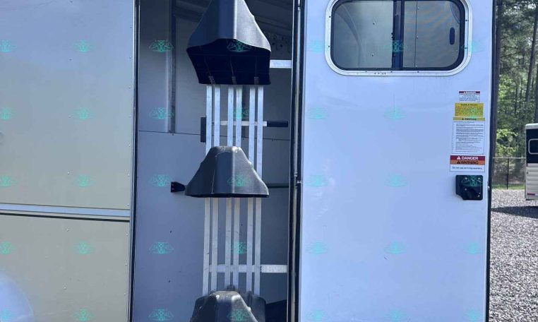 Open silver horse trailer with rear door and ramp down, showing interior stalls on a gravel lot with trees in the background