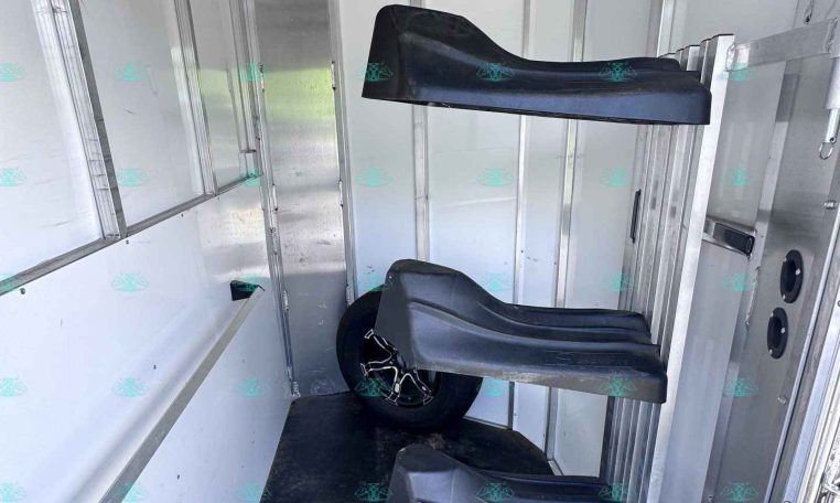 Interior of a metal trailer with two black contoured seats mounted along the wall and a wheel visible toward the back.