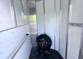 Interior of an aluminum trailer with hooks along the left wall, vertical wall panels, and a spare tire resting near the back on a dark floor.