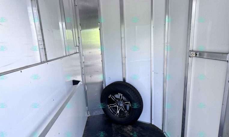 Interior of an aluminum trailer with hooks along the left wall, vertical wall panels, and a spare tire resting near the back on a dark floor.