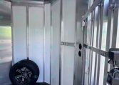 Interior of an aluminum trailer with white wall panels, spare tire resting on the floor, and metal wall framing with a door on the right.