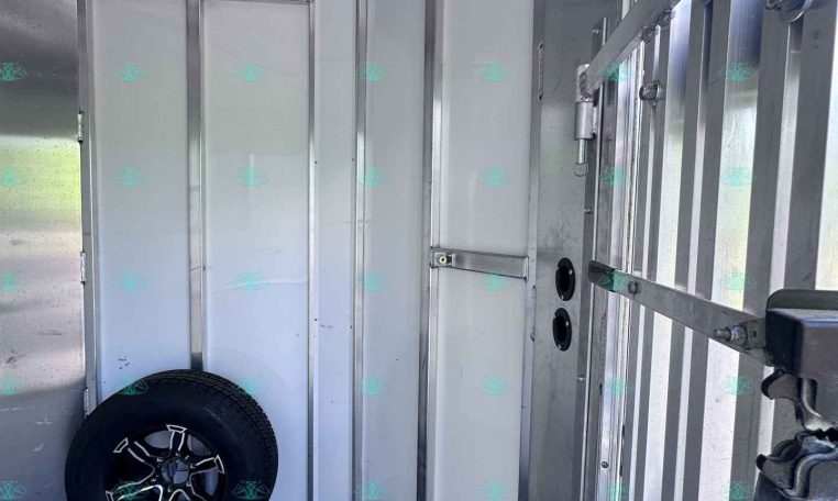 Interior of an aluminum trailer with white wall panels, spare tire resting on the floor, and metal wall framing with a door on the right.