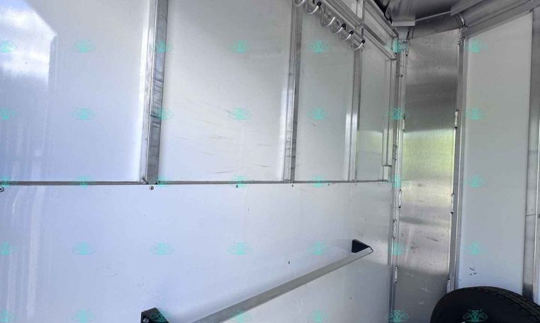 Interior view of a silver metal trailer with a row of hooks along the ceiling and a spare tire in the corner on a dark floor