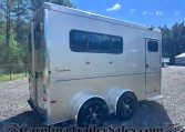 Side view of a silver Sundowner travel trailer with two windows and dual rear wheels, parked on a gravel lot with trees in the background.