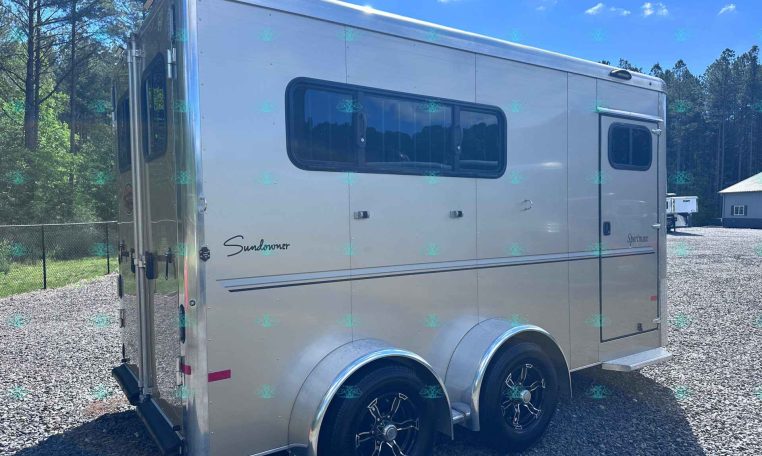 Side view of a silver Sundowner travel trailer with two windows and dual rear wheels, parked on a gravel lot with trees in the background.