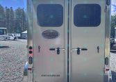 Rear view of a silver double-door trailer with windows, logo plate, and website americatrailersales.com visible on gravel lot.