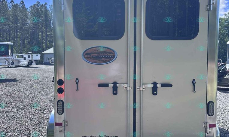 Rear view of a silver double-door trailer with windows, logo plate, and website americatrailersales.com visible on gravel lot.