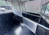 Interior of a metal horse trailer with divided stalls and black padding along the walls. Window bars along the side.