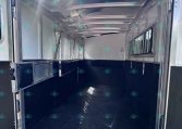 Interior of an empty aluminum horse trailer with side dividers and windows, daylight entering from rear open door, black rubber floor