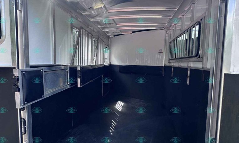 Interior of an empty aluminum horse trailer with side dividers and windows, daylight entering from rear open door, black rubber floor