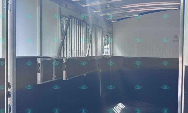 Interior of a horse trailer with partitioned stalls and metal bars, door open to the outside light.
