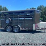 Dark gray horse trailer with side vents and a mounted spare tire on a gravel lot, pine trees in the background.