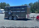 Dark gray horse trailer with side vents and a mounted spare tire on a gravel lot, pine trees in the background.