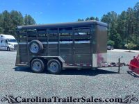 Dark gray horse trailer with side vents and a mounted spare tire on a gravel lot, pine trees in the background.