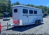 White long horse trailer with side windows sits on a gravel lot, hitch connected, trees in the background.