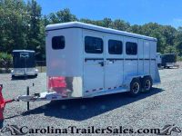 White long horse trailer with side windows sits on a gravel lot, hitch connected, trees in the background.