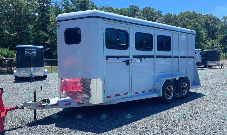 White long horse trailer with side windows sits on a gravel lot, hitch connected, trees in the background.