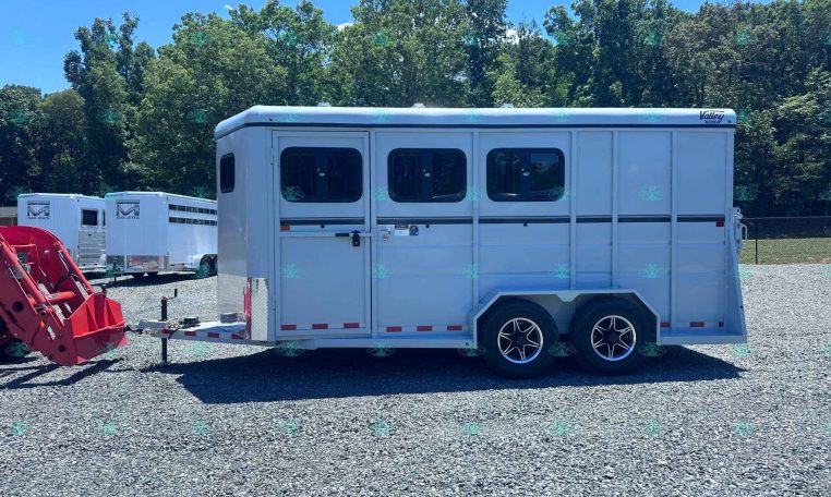 White horse trailer on a gravel lot with trees in the background and other trailers nearby, under a clear blue sky.
