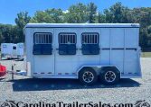 White horse trailer with barred side windows and two axles parked on gravel, trees in the background.