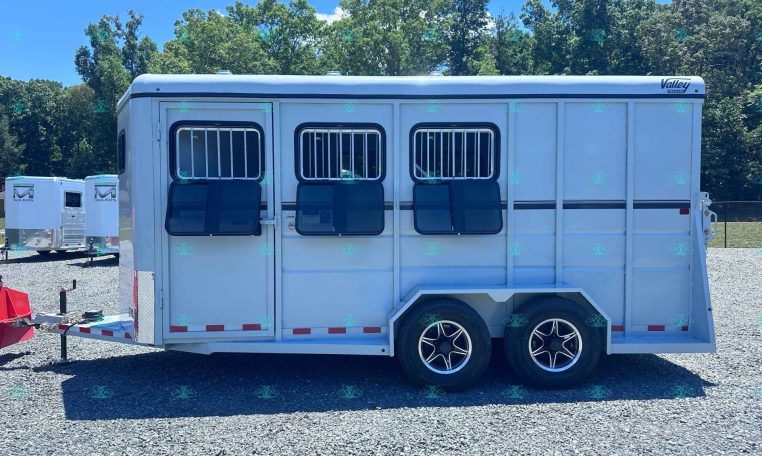 White horse trailer with barred side windows and two axles parked on gravel, trees in the background.