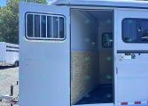 White utility trailer with the side door open, revealing plywood walls and a window barred on the left, parked on gravel under a blue sky with trees in the background.