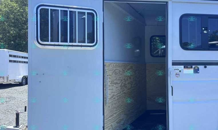 White utility trailer with the side door open, revealing plywood walls and a window barred on the left, parked on gravel under a blue sky with trees in the background.